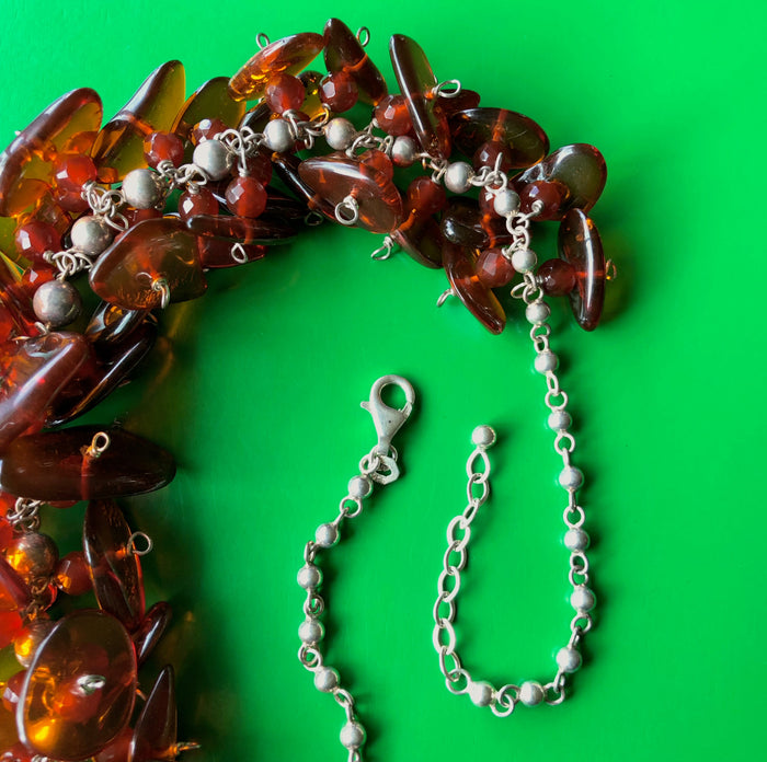 Close up, in-focus, overhead shot of the sterling silver lobster claw clasp of the Cognac Baltic Amber and Faceted Carnelian on Sterling Silver Ball Chain Necklace, laying on a vibrant green wooden surface.