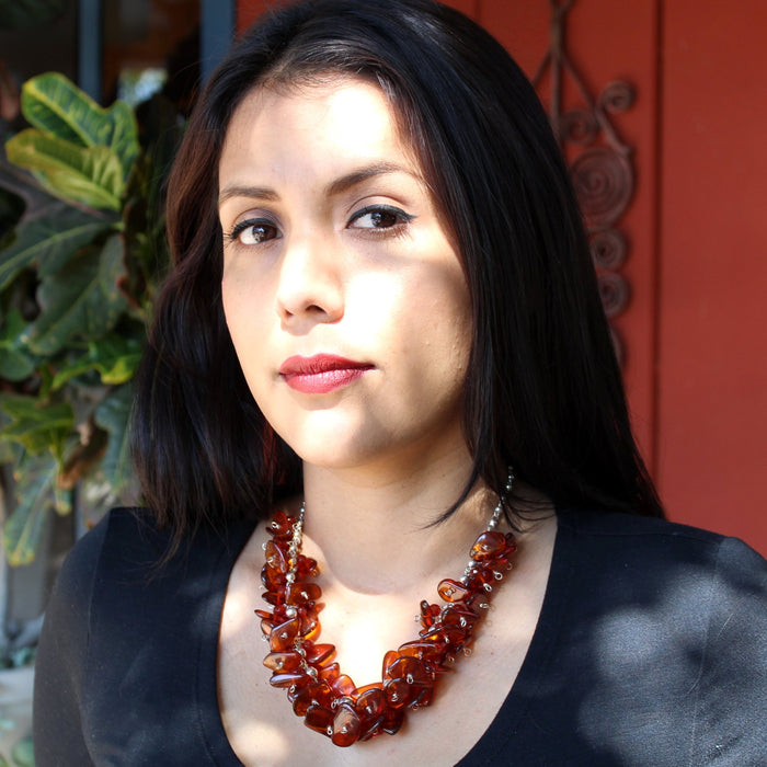 A brunette model with red lipstick, black eyeliner, dark brown eyes in a black long sleeve, scoop neck top wearing the Cognac Baltic Amber and carnelian on Sterling Silver Ball Chain Necklace, sitting outside in front of a potted plant and rust red paneled wall. The necklace sits just below the collar bone, with shadows and sunlight dancing on her face, illuminating the rich golden tones in the amber necklace as well.