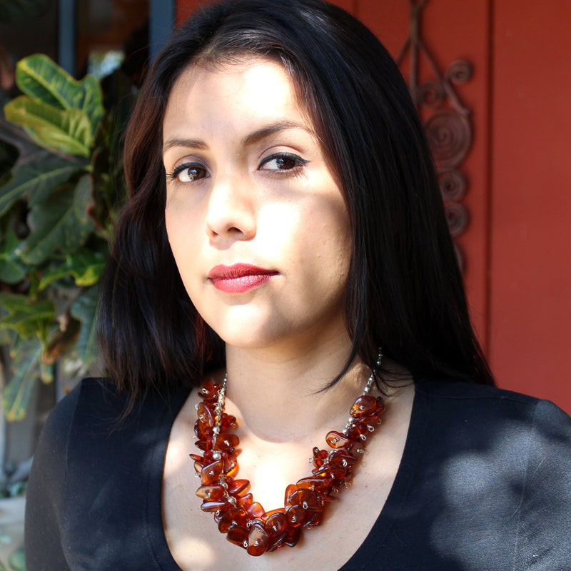 A brunette model with red lipstick, black eyeliner, dark brown eyes in a black long sleeve, scoop neck top wearing the Cognac Baltic Amber and carnelian on Sterling Silver Ball Chain Necklace, sitting outside in front of a potted plant and rust red paneled wall. The necklace sits just below the collar bone, with shadows and sunlight dancing on her face, illuminating the rich golden tones in the amber necklace as well.