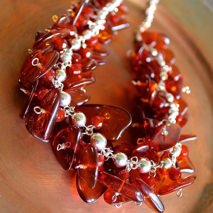 Close up, detailed shot of the Cognac Baltic Amber and Faceted Red Carnelian on Sterling Silver Ball Chain Necklace taken at a 45 degree angle overhead. The beads in the foreground are in focus, showing the detail and minimal texture inside the smooth, flat, oblong amber beads, the bright shine of the silver, and facets of the carnelian beads. The necklace sits on a matte copper plate.