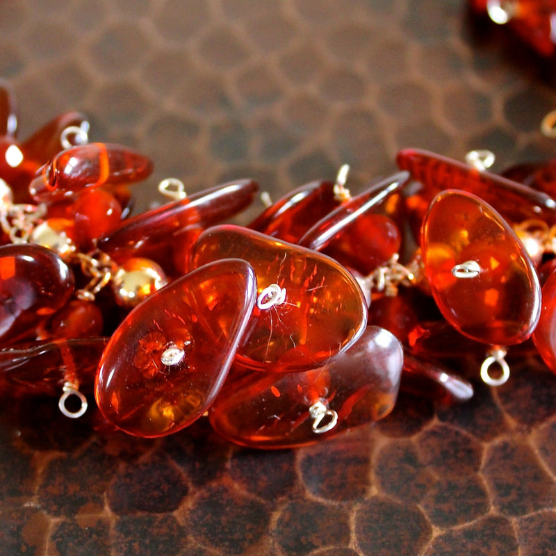 Ultra close up, detailed shot of the Cognac Baltic Amber and Faceted Carnelian on Sterling Silver Ball Chain Necklace taken at a 45 degree angle overhead. The beads in the foreground are in focus, showing the detail and minimal texture inside the smooth, flat, oblong amber beads, and some of the hand wiring througout. The necklace sits on a dark brown hand hammered copper table.
