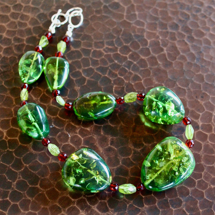 An overhead shot of the Caribbean Green Amber, Red Baltic Amber and Faceted Peridot Bead Necklace, with the entire necklace in focus except for the extender toggle clasp, laying on a dark brown hand hammered copper table. The vibrant green, transluscent amber color shines in natural light, showing off the detailed air bubble textures inside the unique, oblong shaped pieces of amber.