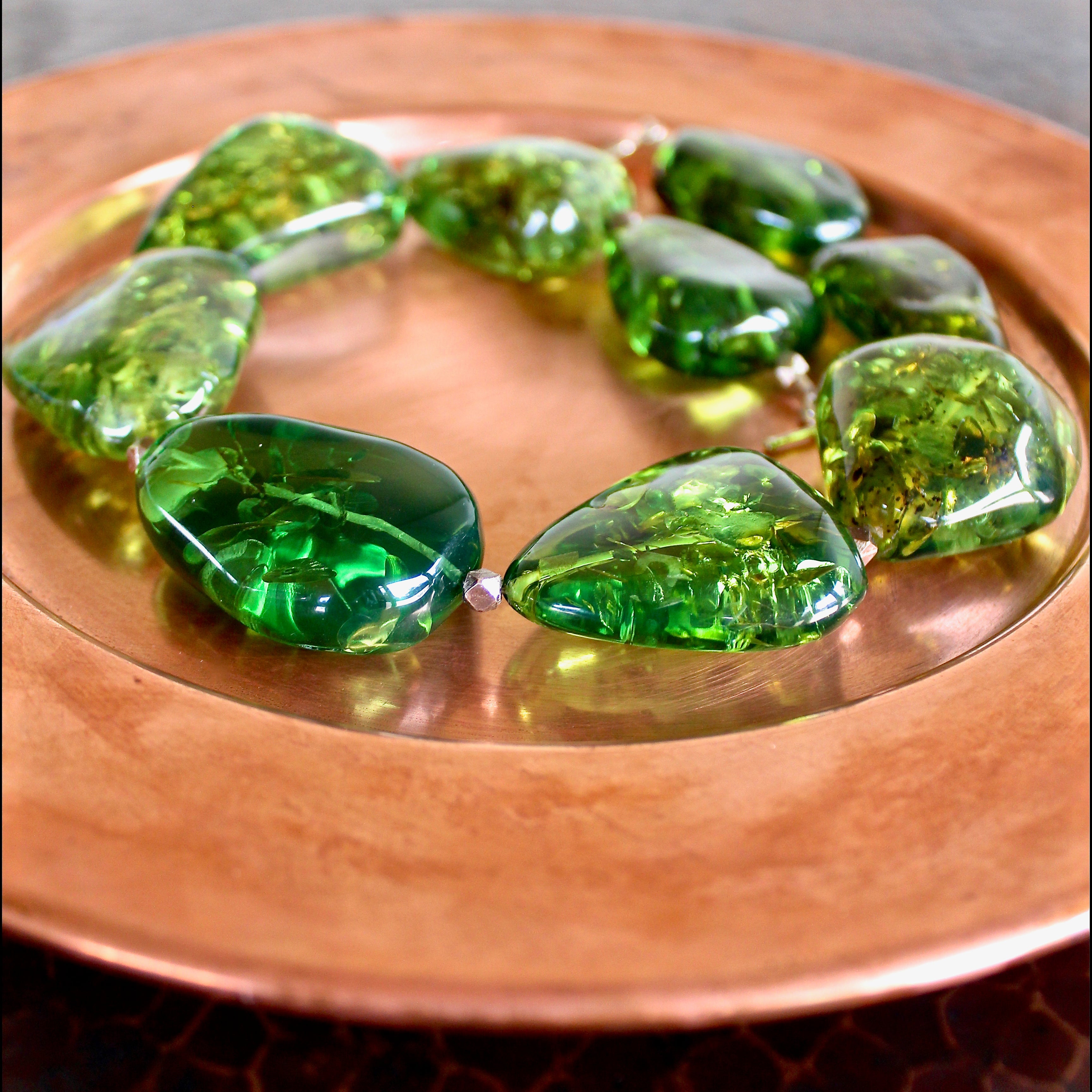 An almost eye level shot of the Caribbean Green Amber and Hill Tribe Silver Necklace laying on a matte copper plate, showing off the thickness and detail of each vibrant green, one of a kind bead.