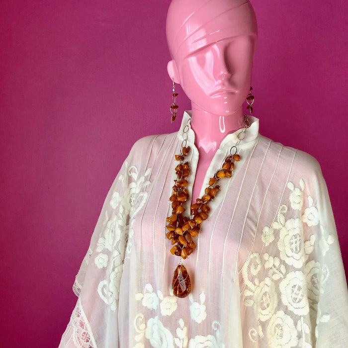 An eyeless pale pink mannequin wears an embroidered, sheer white, Mexican kaftan against a darker pink wall, while modeling Cognac and Butterscotch Baltic Amber Necklace and Earrings Set. The necklace sits at about the mannequin’s sternum and the earrings fall a couple inches above the shoulders.