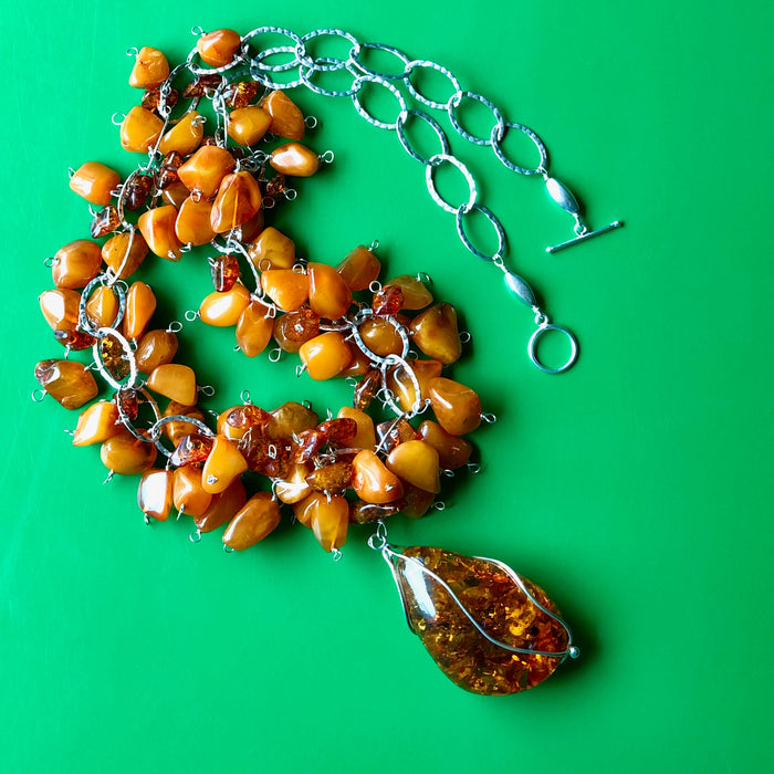 An overhead shot of the Cognac and Butterscotch Baltic Amber Necklace, with the entire necklace in focus, laying on a green wooden surface. The necklace is 28 inches and features Cognac Baltic amber pendant (3 inches) wrapped artfully in sterling silver wire, butterscotch Baltic amber and cognac Baltic amber beads, all on sterling silver chain with a sterling silver clasp