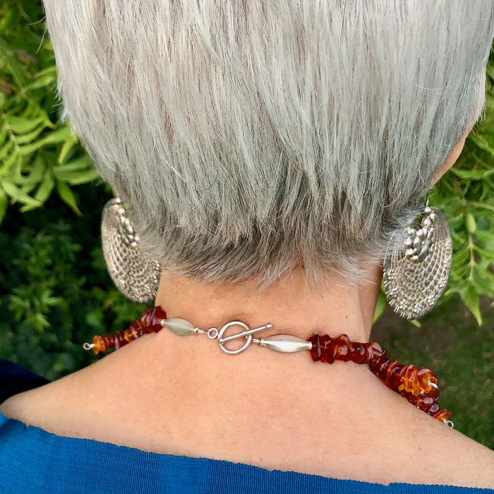 The owner of Joy By Carle, Sylvia, models the Whiskey Baltic Amber Pendant and Baltic Amber Bead Collar Necklace from the back, showing off the sterling silver toggle clasp. Her vibrant turquoise reboso frames the bottom of the shot.