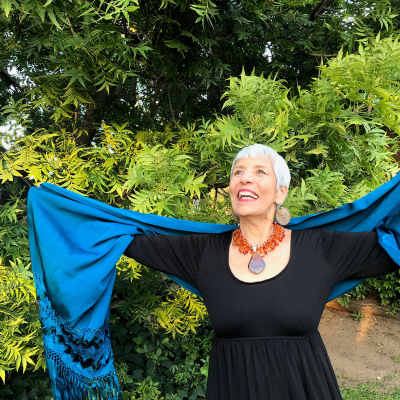 The owner of Joy By Carle, Sylvia, models the Whiskey Baltic Amber Pendant and Baltic Amber Bead Collar Necklace while in a state of jubilation, with arms outstretched upwards and a big, open smile on her face. She is wearing a black, scoop neck, long sleeve, high waisted jersey dress and a vibrant turquoise reboso.