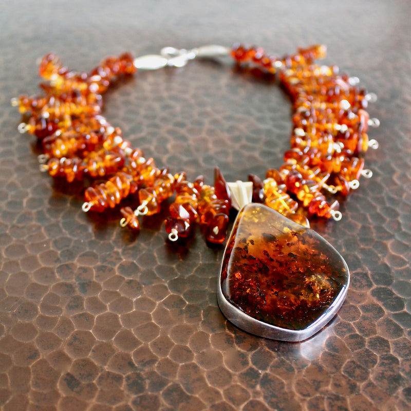 An almost eye level shot of the entire collar necklace laying flat on a hand-hammered copper surface, focusing on the whiskey Baltic amber pendant in sterling silver in the foreground with the whiskey Baltic amber and cognac Baltic amber beaded collar at the back of the frame, becoming more out of focus as your eye moves to the clasp.