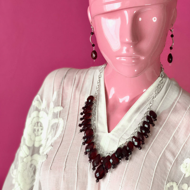 An eyeless pale pink mannequin wears an embroidered, sheer white, Mexican kaftan against a darker pink wall while modeling the Faceted Red Baltic Amber, Red Garnet, Sterling Silver and Hill Tribe Silver Beaded Set, showing off the longer 21.5 inch length of the necklace and 3 inch earrings. The longest part of the necklace sits at about the mannequin’s cleavage.