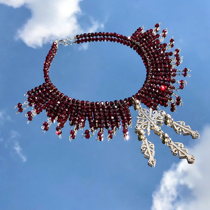 An overhead shot of the Garnet Beads and Oaxacan Sterling Silver Yalalag Cross Collar Necklace, with the entire set in focus. Laying on a vintage Mexican metal framed mirror, the garnets sparkle in the sunlight, and the necklace appears to be floating in the cloudy blue sky. 