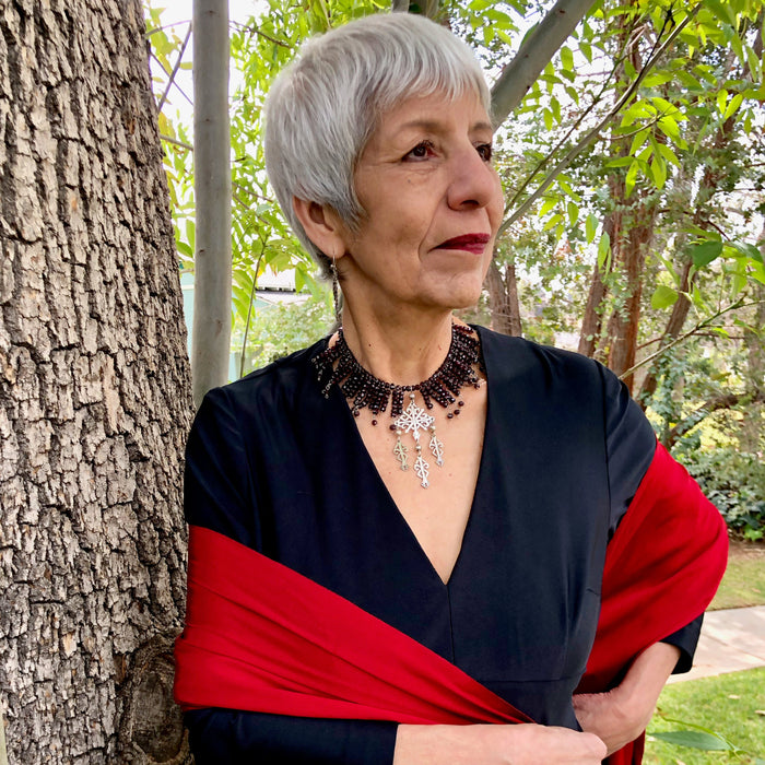 The owner stands next to an ash tree trunk with silvery white hair and red lipstick. She wears a vintage, black, long sleeve, v-neck maxi dress and a vibrant, hand woven reboso, along with the Garnet Beads and Oaxacan Sterling Silver Yalalag Cross Collar Necklace, showing off the collar effect of the design.