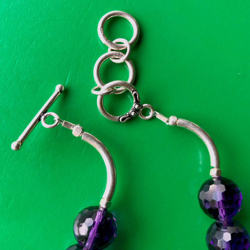 Ultra close up, detailed shot of the sterling silver extender toggle clasp with a detailed view of some of the circle shape, faceted amethyst beads. The necklace is laying on a bright green wooden surface.Ultra close up, detailed shot of the sterling silver extender toggle clasp with a detailed view of some of the circle shape, faceted amethyst beads. The necklace is laying on a bright green wooden surface.