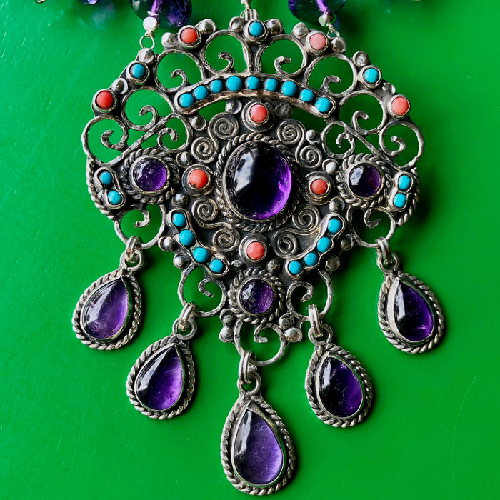  Close up shot of the Matilde Poulat-Style Pendant with Hill Tribe Pure Silver and Faceted Amethyst Beaded Necklace, spotlighting the magnificent pendant of coral, turquoise and larger amethyst cabochons with intricate sterling silver work. The necklace is laying on a bright green wooden surface.