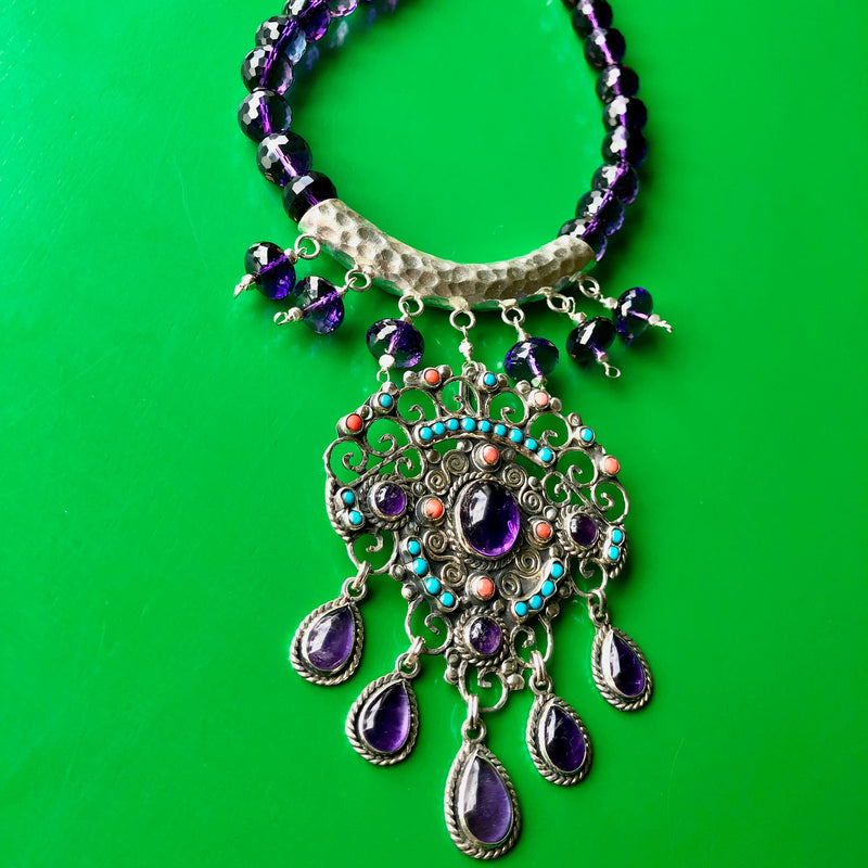 Close up shot of the Matilde Poulat-Style Pendant with Hill Tribe Pure Silver and Faceted Amethyst Beaded Necklace, spotlighting the magnificent pendant of coral, turquoise and larger amethyst cabochons along with the bottom half of the necklace including the hand hammered Hill Tribe pure silver pendant with 6 hanging amethyst beads. The necklace is laying on a bright green wooden surface.