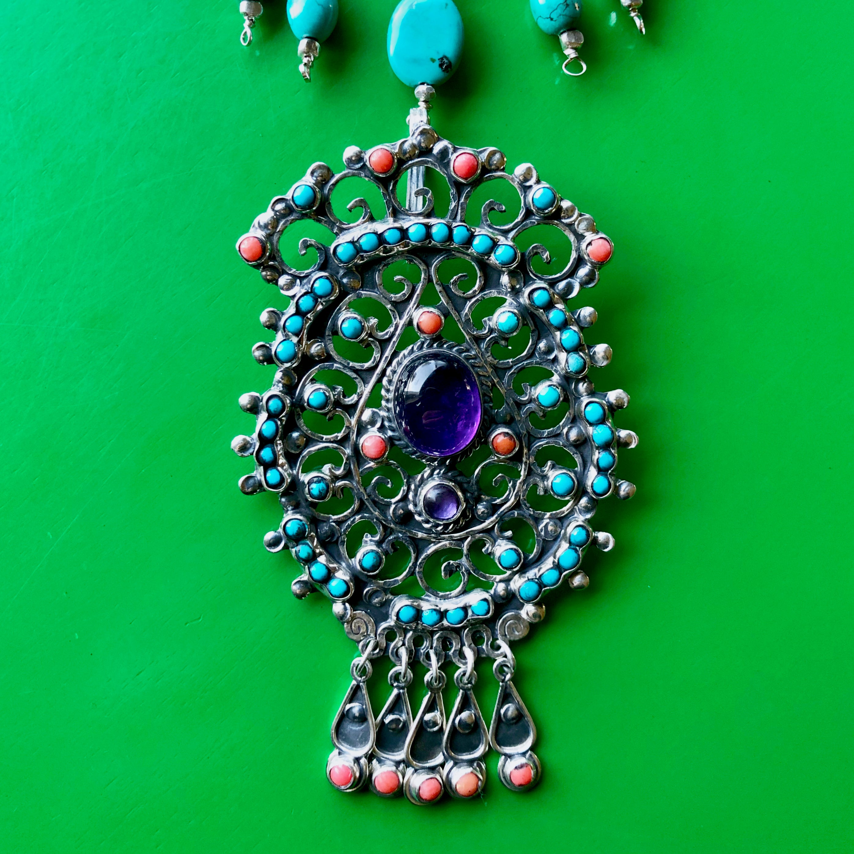 Close up shot of the Matilde Poulat-Style Pendant with Hill Tribe Pure Silver nuggets and turquoise beads in the frame, spotlighting the magnificent pendant of coral, turquoise and larger amethyst cabochons with intricate sterling silver work. The piece is laying on a bright green wooden surface.
