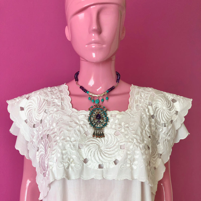 A pink mannequin wears an embroidered white huipil in front of a darker pink wall while modeling the Matilde Poulat Style Pendant on Amethyst and Fine Turqoise Beaded Necklace. The necklace sits just above the collarbone and longest part of the pendant reaches at about the center of the chest.