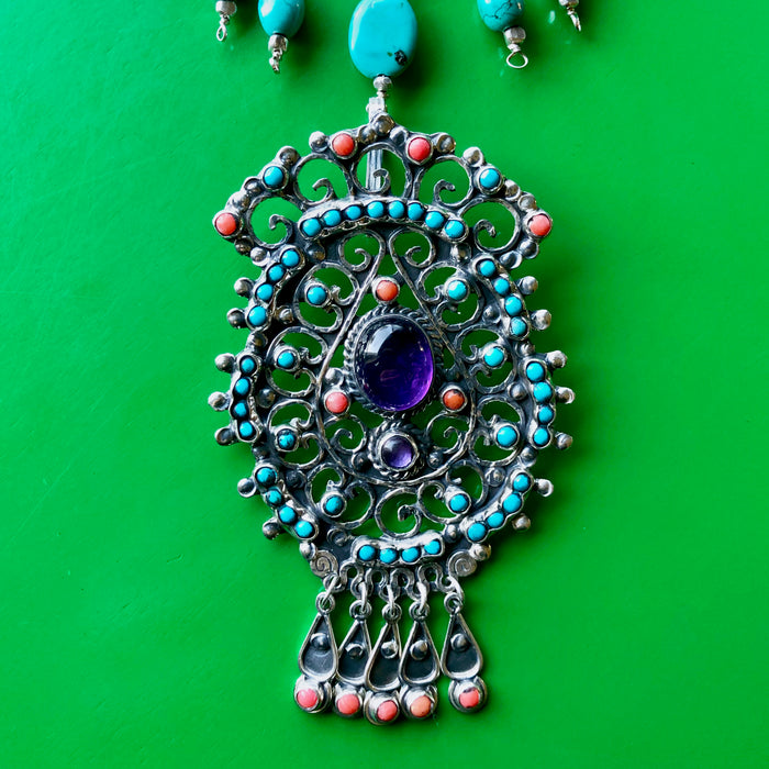 Close up shot of the Matilde Poulat-Style Pendant with Hill Tribe Pure Silver nuggets and turquoise beads in the frame, spotlighting the magnificent pendant of coral, turquoise and larger amethyst cabochons with intricate sterling silver work. The piece is laying on a bright green wooden surface.
