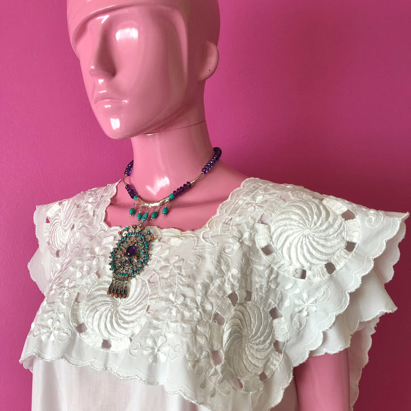 Pink mannequin at three quarter view in front of a darker pink wall wearing a white embroidered huipil from the Yucatan and the handmade beaded necklace with the Matilde Poulat inspired pendant that anchors a large, hand-hammered Hill Tribe pure (.999) silver pendant, Hill Tribe pure silver spacers, and faceted amethyst and turquoise beads. The necklace is sitting at the collarbone with the pendant reaching down to about the center of the chest.