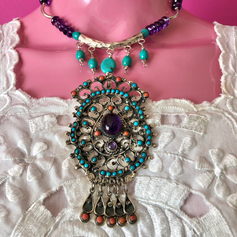 Pink mannequin at front facing view wearing a white embroidered huipil from the Yucatan. Sitting just at the base of the neck, the handmade beaded necklace with fine turquoise beads hanging from the hand hammered Hill Tribe pure silver pendant is anchored by the sterling silver Matilde Poulat inspired pendant with amethyst cabachon, turquoise and coral. The image is cropped in close, in focus with lots of detail.