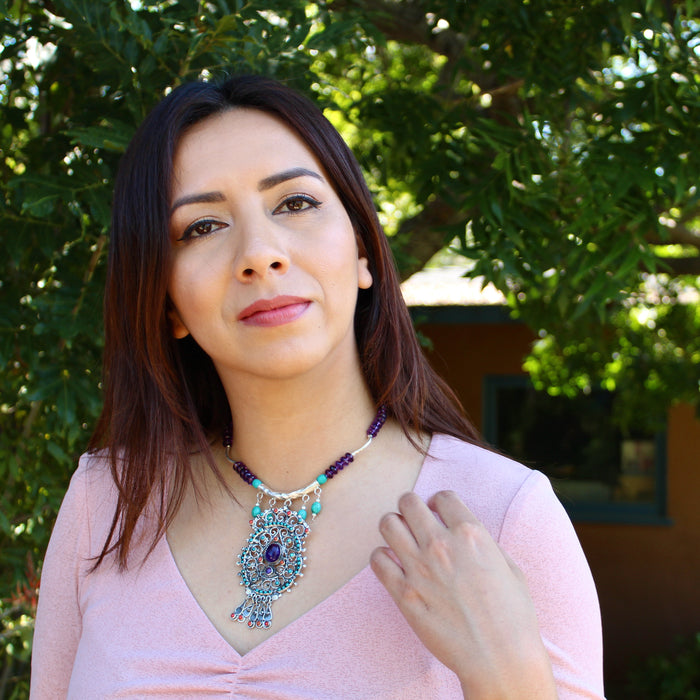 The model with dark, auburn tinted hair stands front facing with a large tree in the background. She is in black eyeliner and reddish pink lipstick, wearing a soft pink v-neck top along with the Amethyst Turquoise Matilde Poulat Style Pendant Beaded Necklace that sits right at the base of her neck with the pendant cascading down to the center of her chest.