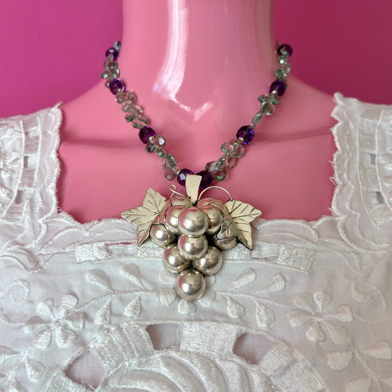 Pink mannequin at front facing view wearing a white embroidered huipil from the Yucatan. The handmade beaded necklace with fine green and purple amethyst is anchored by the vintage, sterling silver grape cluster + leaves pendant. The image is cropped in close, in focus with lots of detail including the sculptural roundness of grapes and line work in the two grape leaves.