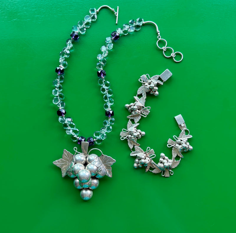 Overhead shot showing, in their entirety, the vintage sterling silver grape clusters bracelet and necklace with matching grape cluster pendant, faceted green and purple amethyst beads, Hill Tribe pure silver spacers and nuggets, and sterling silver extender toggle clasp. The set is laying on a green wooden surface.
