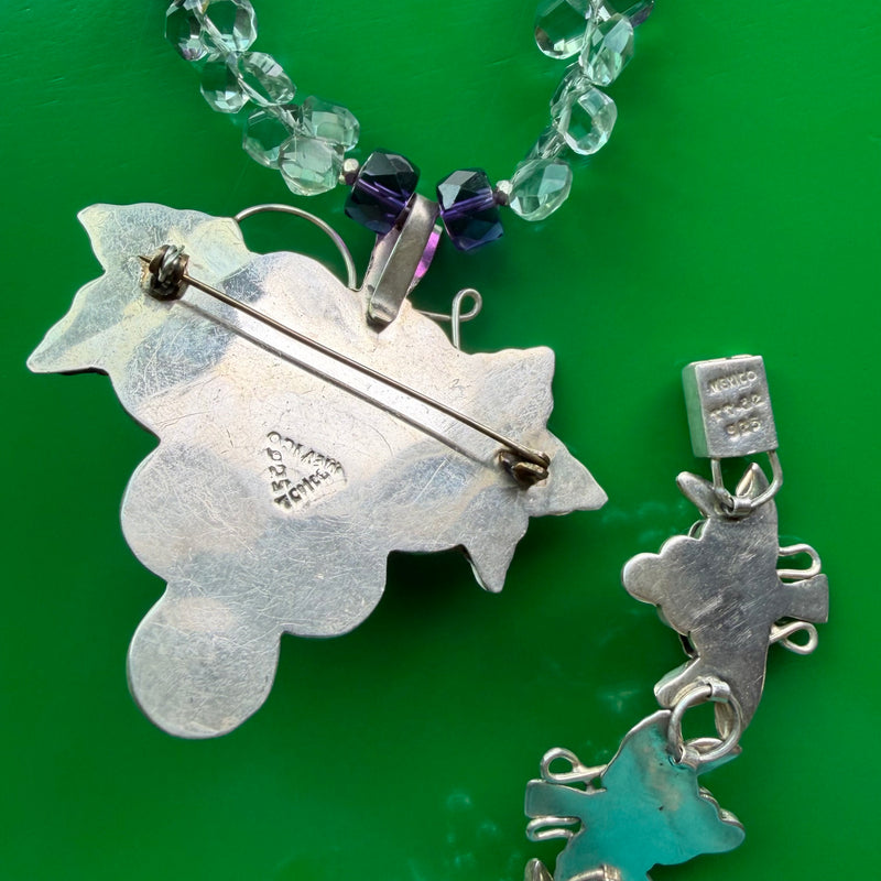 Closeup shot of the backside of the pendant and the back of the box clasp of the bracelet - laying side by side on a green wood surface, showing the 925 stamps on both indicating the two pieces are sterling silver.