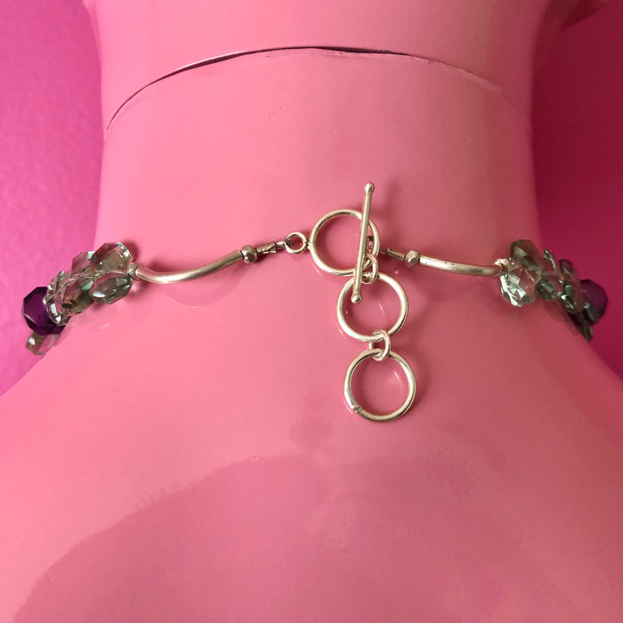 Close up shot of the extender toggle clasp and a small portion of the handmade beaded necklace, including the faceted green and purple amethyst beads and large Hill Tribe pure silver spacers and nuggets. Shown on a pink mannequin facing a darker pink wall.