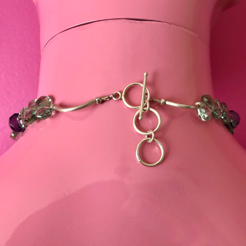 Close up shot of the extender toggle clasp and a small portion of the handmade beaded necklace, including the faceted green and purple amethyst beads and large Hill Tribe pure silver spacers and nuggets. Shown on a pink mannequin facing a darker pink wall.