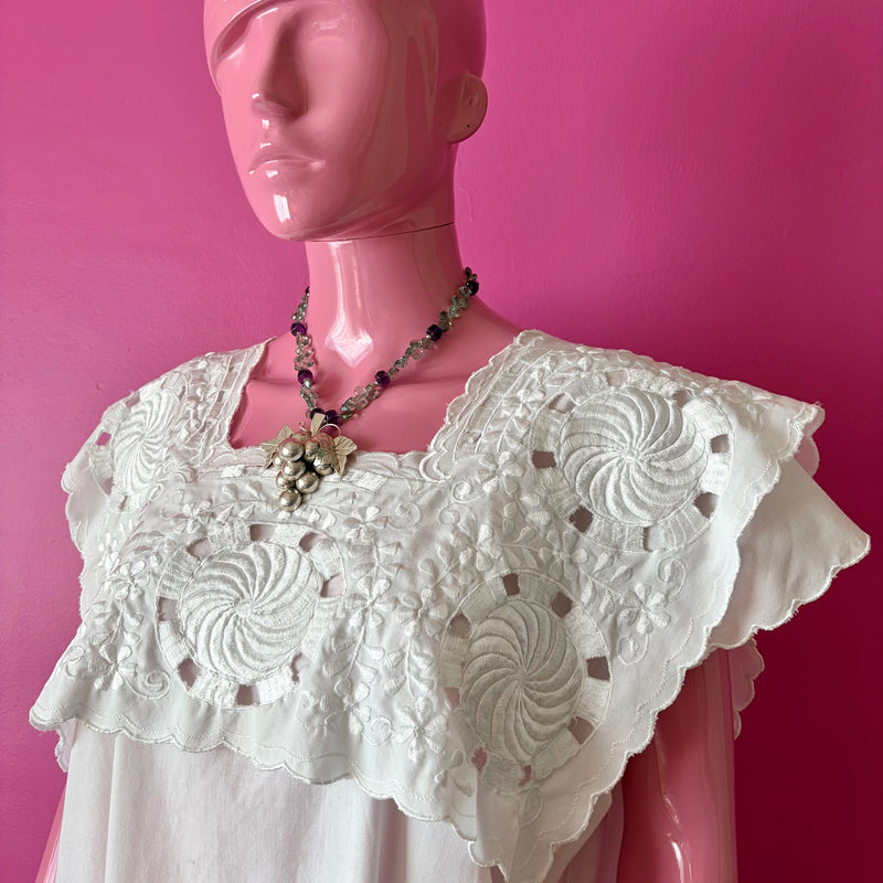 Pink mannequin at three quarter view in front of a darker pink wall wearing a white embroidered huipil from the Yucatan and the handmade beaded necklace with the Vintage Sterling Silver Grape Pendant with Amethyst Beads. The necklace falls at about the collarbone with the pendant reaching down to just above the breast of the mannequin.