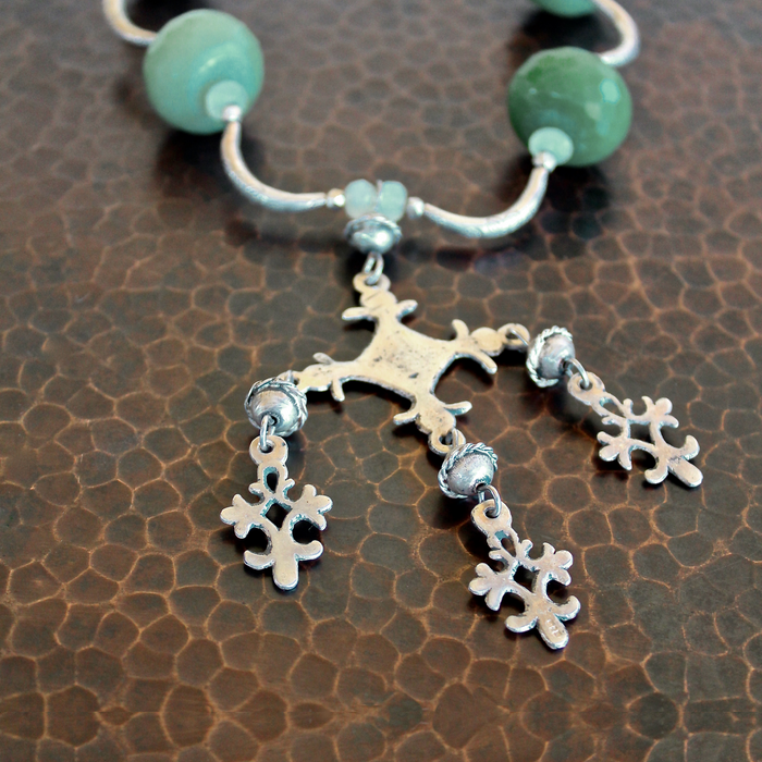 Close up, cropped, detailed overhead shot of the Yalalag pendant necklace with green jade beads, and Hill Tribe pure silver spacers laying on a dark brown, hand hammered copper surface. The image is cropped showing only the pendant in focus, and about half of the necklace fades into the background towards the top of the frame. 