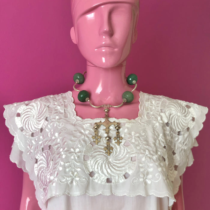 Pink mannequin facing forward in front of a darker pink wall wearing a white embroidered huipil from the Yucatan and the handmade beaded necklace with vintage sterling silver Yalalag cross pendant, large faceted jade beads, small spring jade beads, large Hill Tribe pure silver spacers and nuggets, sterling silver toggle clasp.