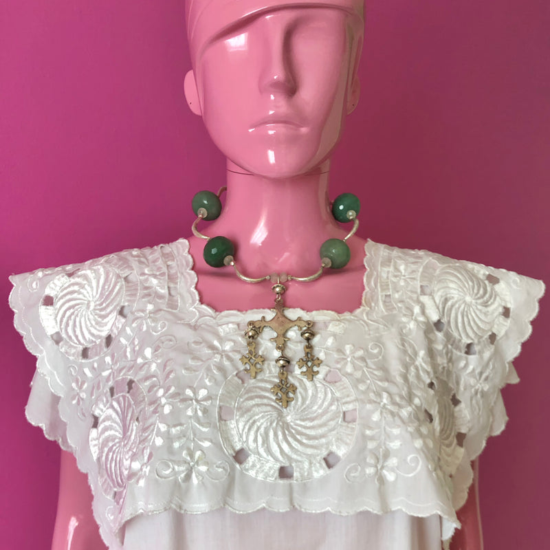 Pink mannequin facing forward in front of a darker pink wall wearing a white embroidered huipil from the Yucatan and the handmade beaded necklace with vintage sterling silver Yalalag cross pendant, large faceted jade beads, small spring jade beads, large Hill Tribe pure silver spacers and nuggets, sterling silver toggle clasp.
