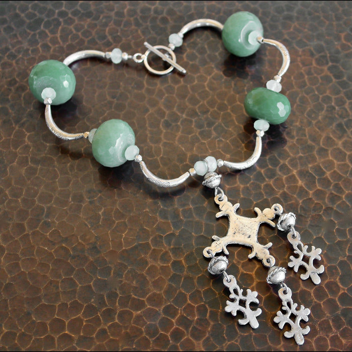 Overhead shot showing the full necklace with vintage sterling silver Yalalag cross pendant, large faceted jade beads, small spring jade beads, large Hill Tribe pure silver spacers and nuggets, sterling silver toggle clasp. The piece is laying on a dark brown, hand hammered copper surface.