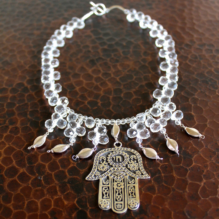 Overhead shot showing the full necklace with the intricate, sterling silver Hamsa (Hand of Fatima) pendant,  faceted rock crystal and sterling beads, large Hill Tribe pure silver spacers and nuggets, and sterling silver toggle clasp. The piece is laying on a dark brown, hand hammered copper surface.