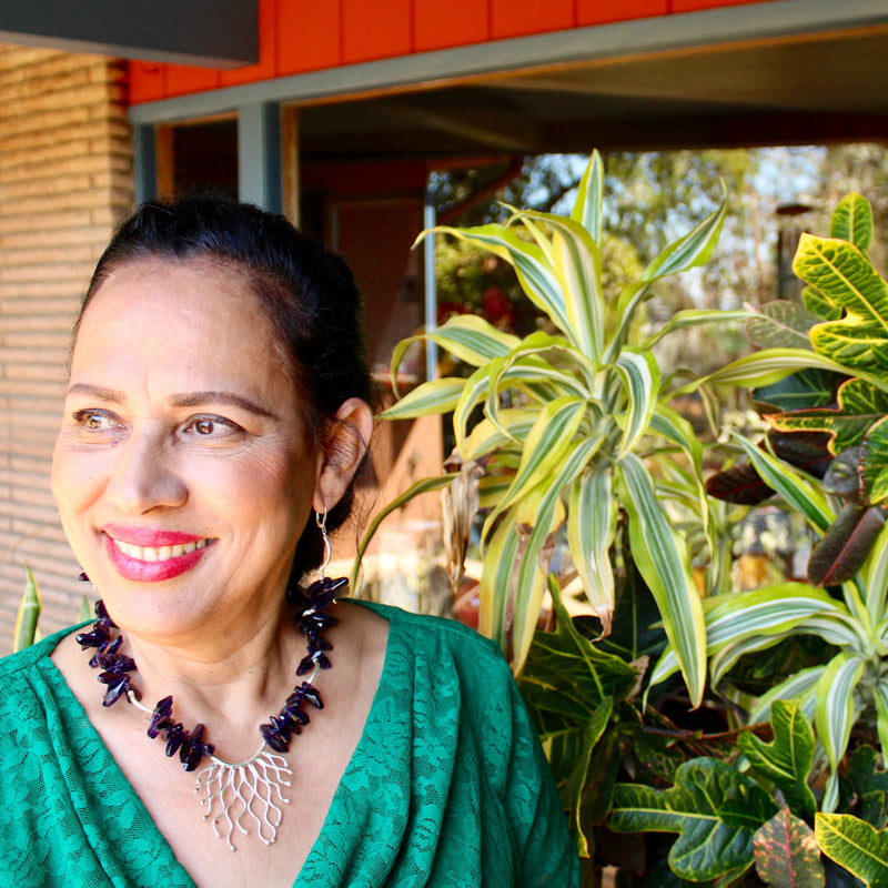 A woman sitting by a window with a green lace blouse and bright smile. She is wearing a necklace and earrings with deep purple amethyst beads and hill tribe silver components, including an abstract pendant in the center. Plants and a midcentury building are in the background.