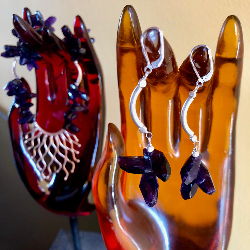 Amethyst earrings displayed on an amber colored hand shape stand with the matching necklace from the set on another red colored hand shaped stand in the blurred background.