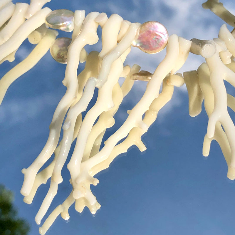Ultra close up, detailed shot of the the White Branch Coral and Coin Pearls Necklace taken at a 45 degree angle overhead. Shows the detail and shape of the coral branch beads, as well as the iridescent sheen on the coin pearl beads. The necklace is laying on a vintage Mexican metal framed mirror, the necklace appears to be floating in the cloudy blue sky. 