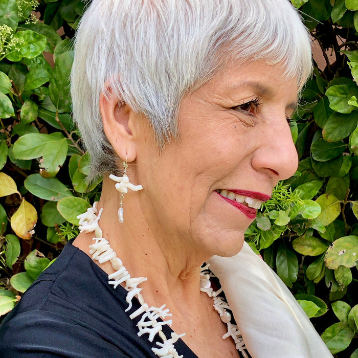 Profile shot of the smiling owner stands in front of green foliage with silver white hair and red lipstick wears a vintage, black, long sleeve, v-neck maxi dress and a cream colored, hand woven reboso, along with the White Branch Coral and Coin Pearls Necklace and Earring Set, showing off the 2.5 inch pearl and white coral dangle earrings.