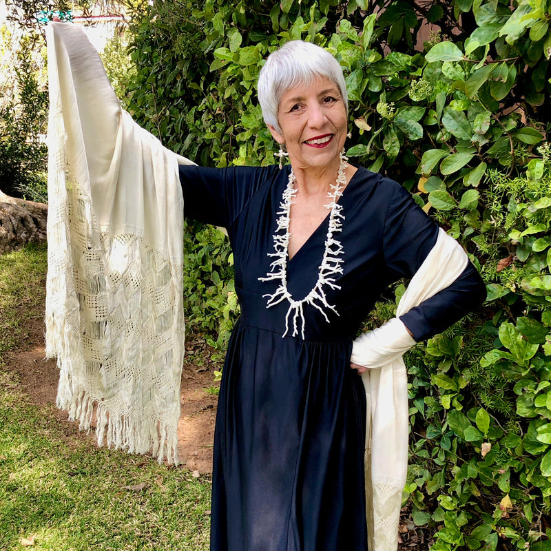 The smiling owner stands in front of green foliage with silver white hair and red lipstick wears a vintage, black, long sleeve, v-neck maxi dress and a cream colored, hand woven reboso, along with the White Branch Coral and Coin Pearls Necklace and Earring Set, showing off the 35 inch necklace that sits at her belly at its longest point.