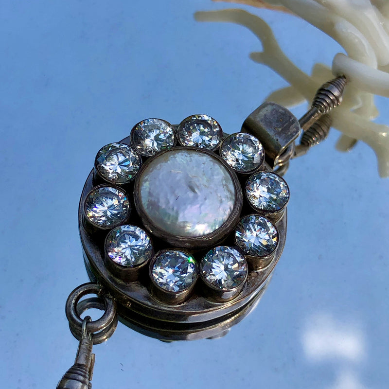 Ultra close up, detailed shot of the Sterling Silver Clasp with Zircons and Pearl clasp, taken at a 45 degree angle overhead. Shows ten sparkling circle shape zircons surrounding one larger circle shaped, lightly textured pearl. The necklace is laying on a vintage Mexican metal framed mirror, the necklace appears to be floating in the cloudy blue sky. 