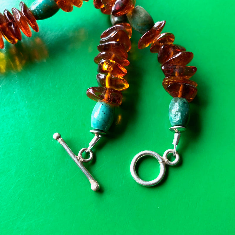 Close up shot of the sterling silver toggle clasp and the amber and turquoise beads on the Tibetan Copal Pendant, Cognac Baltic Amber, and Turquoise Necklace laying on a vibrant green wooden surface.