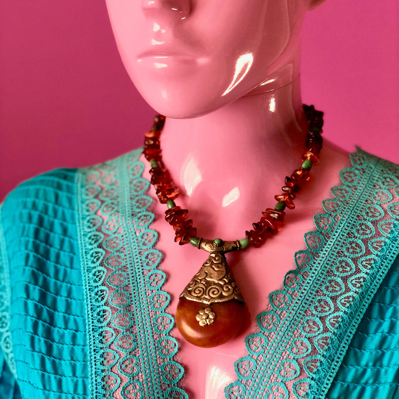 Close-up shot of an eyeless pale pink mannequin modeling the Tibetan Copal Pendant, Cognac Baltic Amber, and Turquoise Necklace while wearing a bright turquoise lace and pin tucked cotton dress with a deep v-neck that allows the necklace to fall flatteringly on the decolletage. The bottom of the pendants sits right at the middle of the mannequin’s chest. 