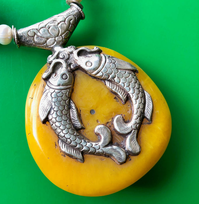 Close up shot of the Tibetan Double-Sided Yellow Copal Pendant with Carp and Dragon, Pearls, Large Copal Beads Necklace, showing off the other side of the incredible, opaque, creamy yellow copal pendant in great detail, including the intricate silver double carp design on the pendant. The necklace is laying on a bright green wooden surface.