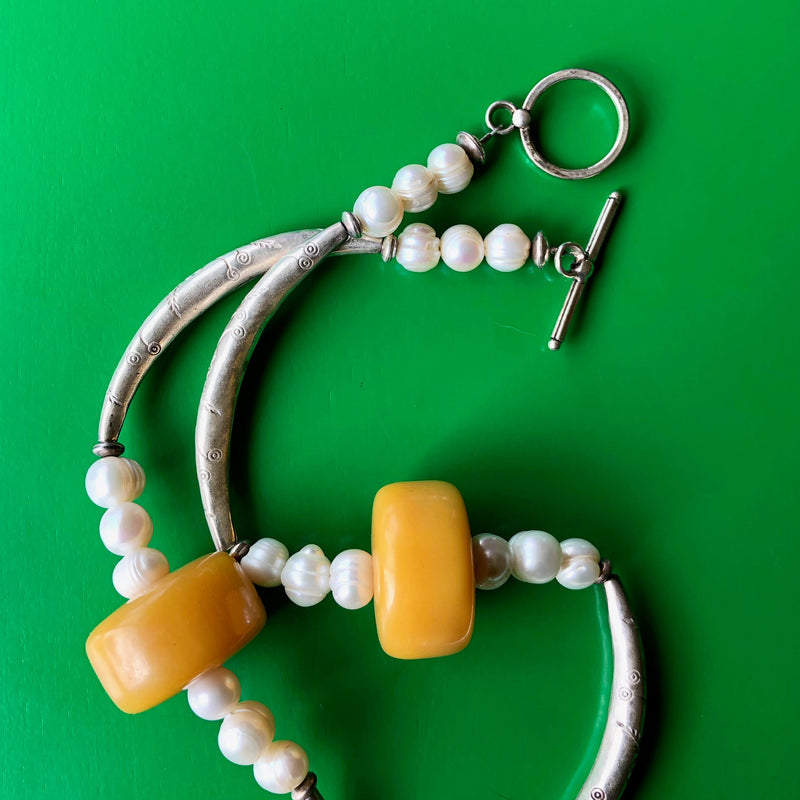 Close up, in-focus, overhead shot of the sterling silver toggle clasp of the Tibetan Double-Sided Yellow Copal Pendant with Carp and Dragon, Pearls, Large Copal Beads Necklace, including two of the vintage copal cylinder beads, along with some of the pearls and Hill Tribe pure silver spacers. Necklace is laying on a vibrant green wooden surface.