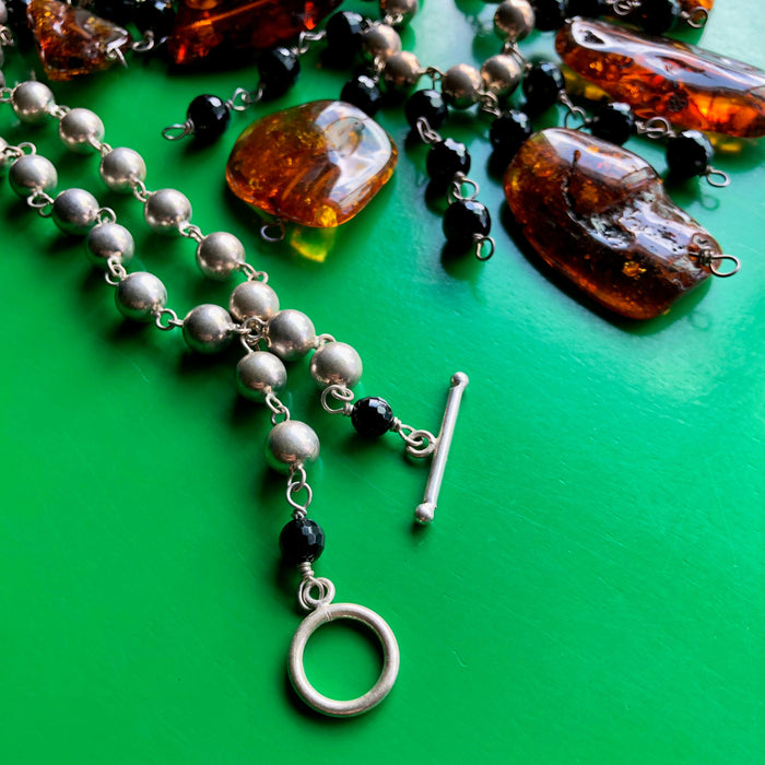 Close up, in-focus shot of the sterling silver toggle clasp of the Whiskey Baltic Amber and Faceted Onyx Beads on Sterling Silver Ball Chain Necklace with the rest of the necklace receding into the background, laying on a vibrant green wooden surface.