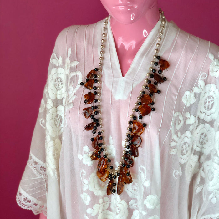 An eyeless pale pink mannequin wears an embroidered, sheer white, Mexican kaftan against a darker pink wall while modeling the Whiskey Baltic Amber and Faceted Onyx Beads on Sterling Silver Ball Chain Necklace, showing off the longer 36 inch length of the necklace. The longest part of the necklace reaches the mannequin’s belly.