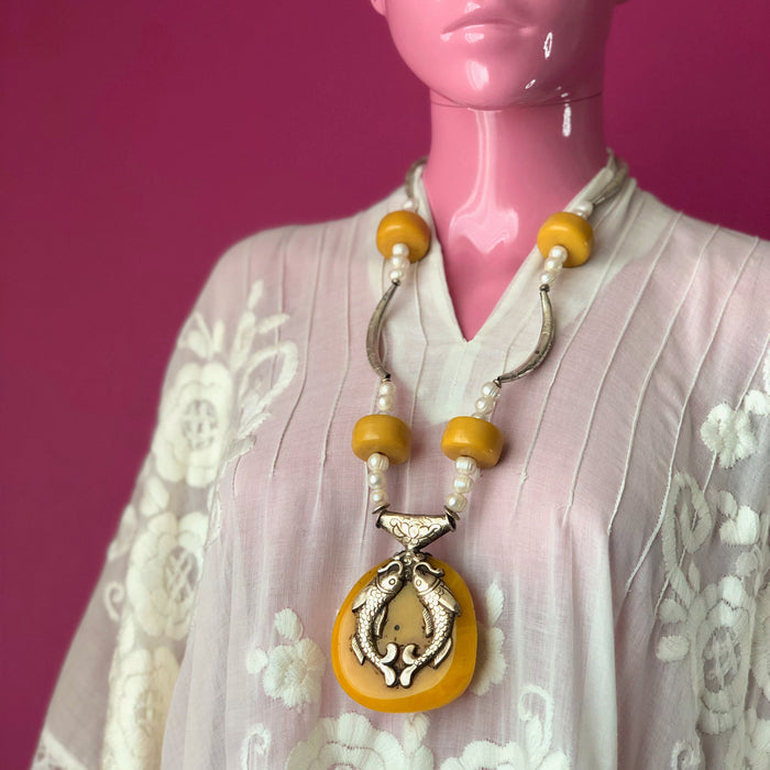 A pale pink mannequin wears an embroidered, sheer white, Mexican kaftan against a darker pink wall while modeling the Tibetan Double-Sided Yellow Copal Pendant with Carp and Dragon, Pearls, Large Copal Beads Necklace, showing off the longer 29 inch length of the necklace and the carp side of the pendant. The longest part of the necklace reaches the mannequin’s sternum.