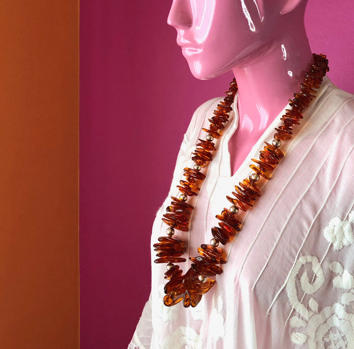 An eyeless pale pink mannequin wears an embroidered, sheer white, Mexican kaftan against the corner of a darker pink wall and a pale orange wall, while modeling the Whiskey Baltic Amber with Hill Tribe Silver Necklace, showing off the translucence of the amber and the luminescence of the pure Hill Tribe silver hand hammered beads. The longest part of the necklace sits at about the mannequin’s sternum.