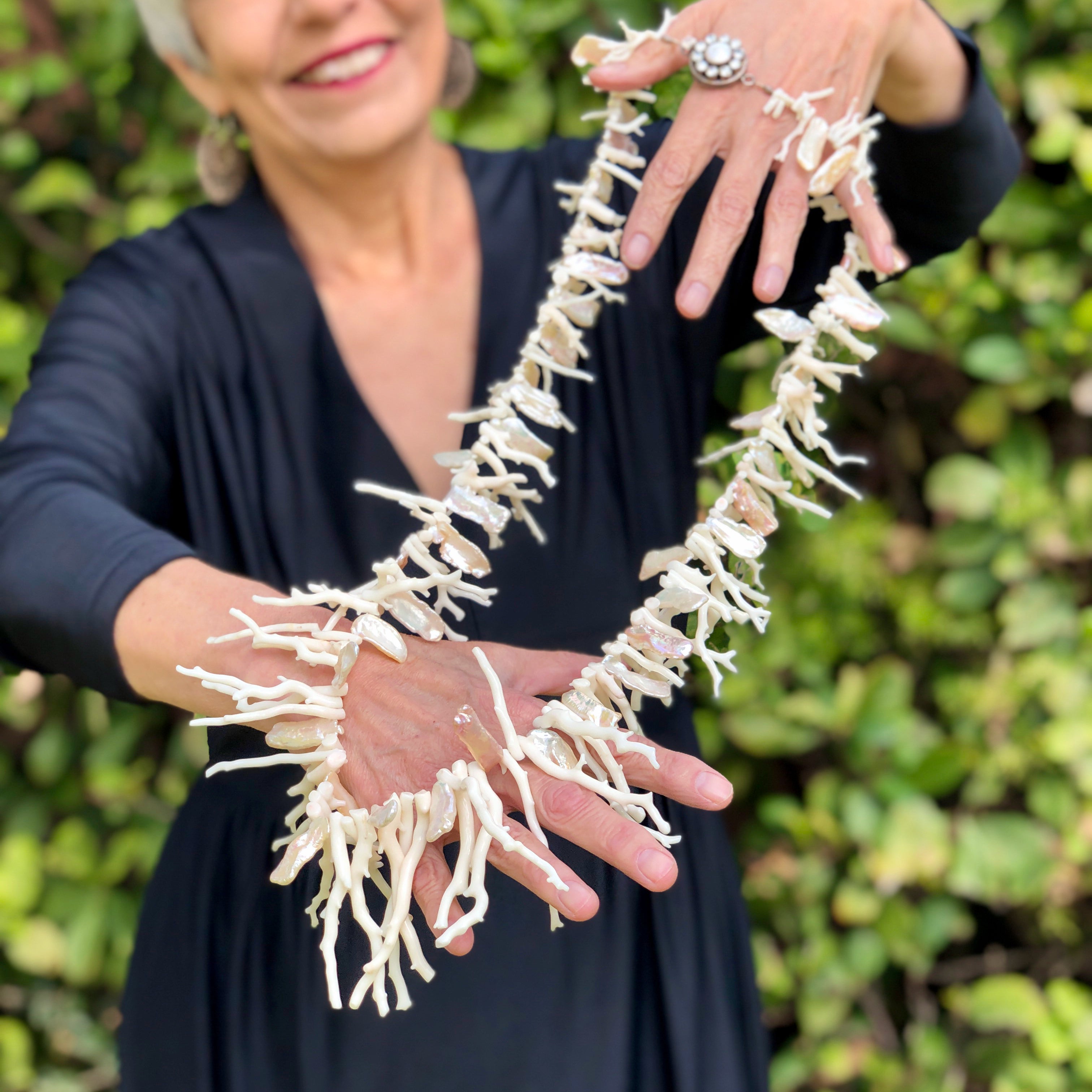 The smiling model stands in front of green foliage with silver white hair and red lipstick wearing a vintage, black, long sleeve, v-neck maxi dress holding up in two hands with arms extended forward, the White Branch Coral, Keshi Pearls, Sterling Silver Clasp with Zircons and Pearl Necklace, showing off the 36 inch necklace in all its glory.